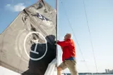 A person stands on a sailboat at the dock, using a tape measure to take accurate measurements along the mast