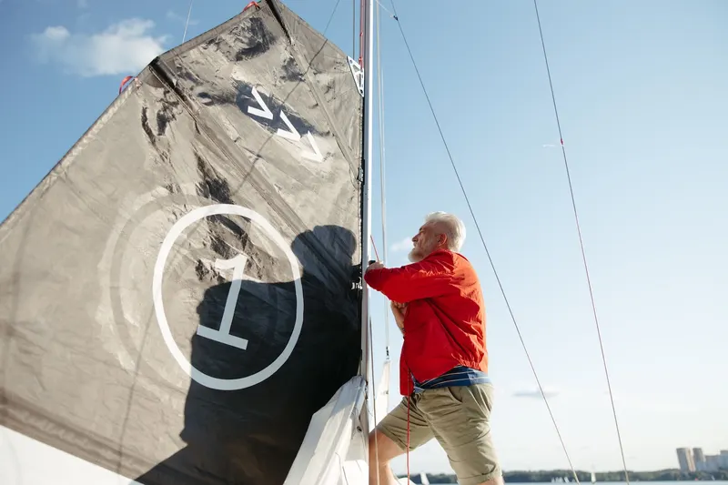A person stands on a sailboat at the dock, using a tape measure to take accurate measurements along the mast
