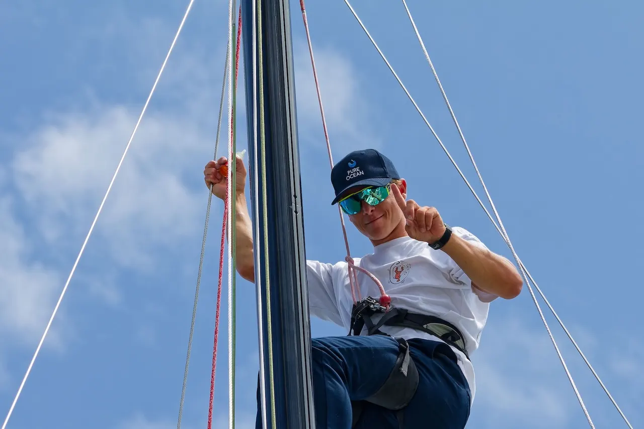 A person in a climbing harness is positioned at the top of a sailboat mast on a clear day